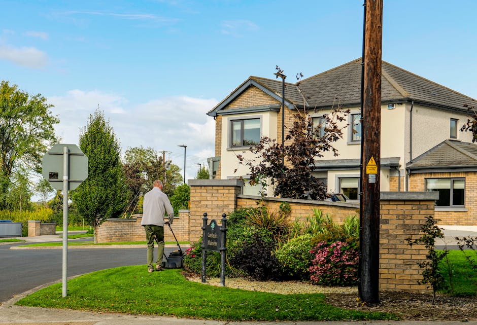 A close-up view of a garden with neatly cut, lush green grass and well-maintained flower beds containing a variety of plants, including small shrubs and flowering perennials. A paved pathway weaves th