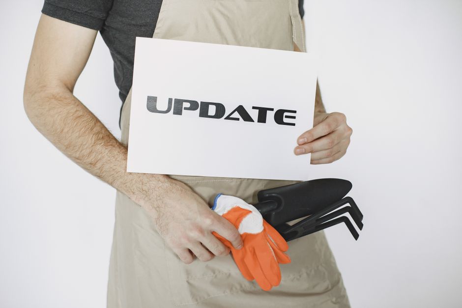 A person wearing a beige gardening apron and a dark grey t-shirt, holding a white sign with the word 'UPDATE' in bold black letters, stands against a plain white background. In one hand, they hold a p